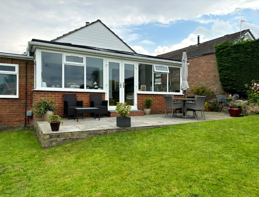A conservatory extension with a new Supalite tiled roof replacement. The conservatory has white frames is attached to a red brick bungalow, viewed from a green lawn. 