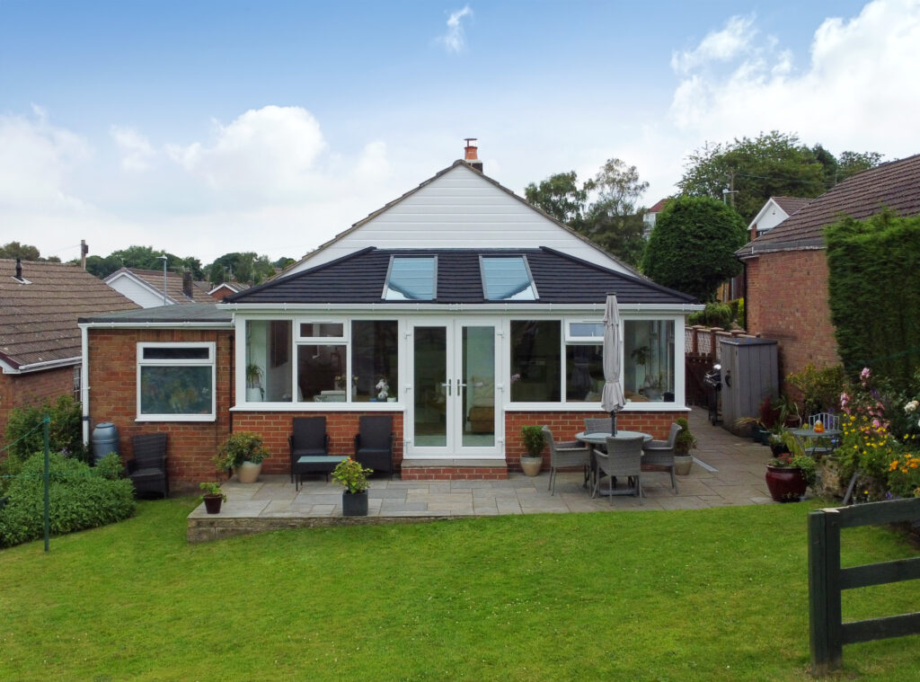 A conservatory with a dark tiled Supalite roof and SKyvista skylights is attached to a red brick bungalow. The conservatory has white-framed windows and doors.